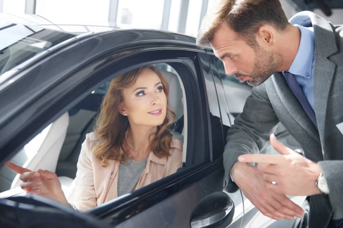 A car salesperson gestures animatedly while discussing features with a potential buyer inside a modern car showroom.
