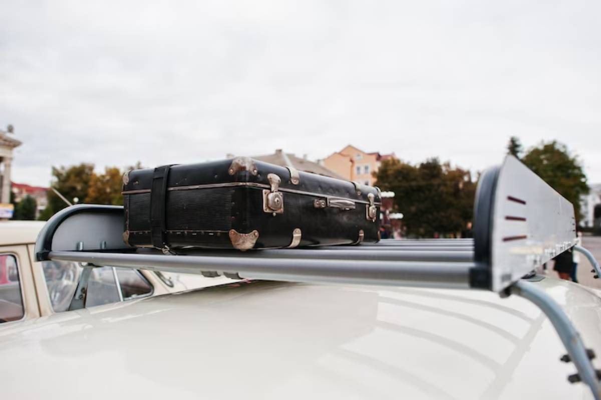 A vintage black suitcase rests on the roof rack of a car, with buildings and trees in the background under a cloudy sky.