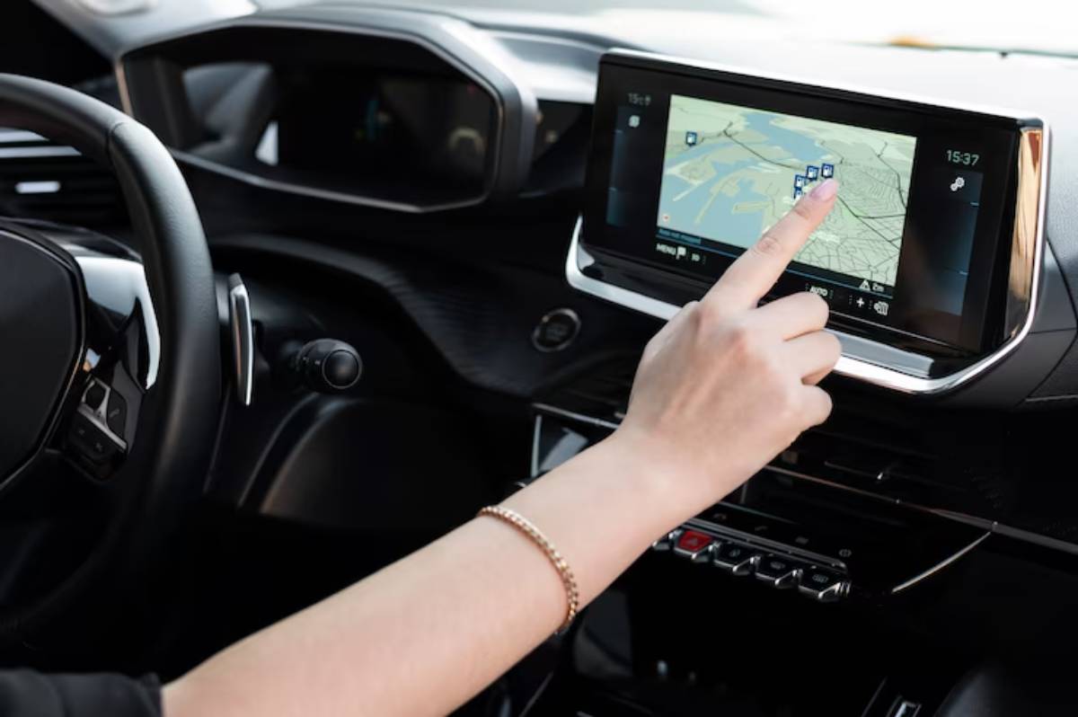 A hand touches a car's touchscreen display showing a map and navigation prompts, with a steering wheel partially visible.