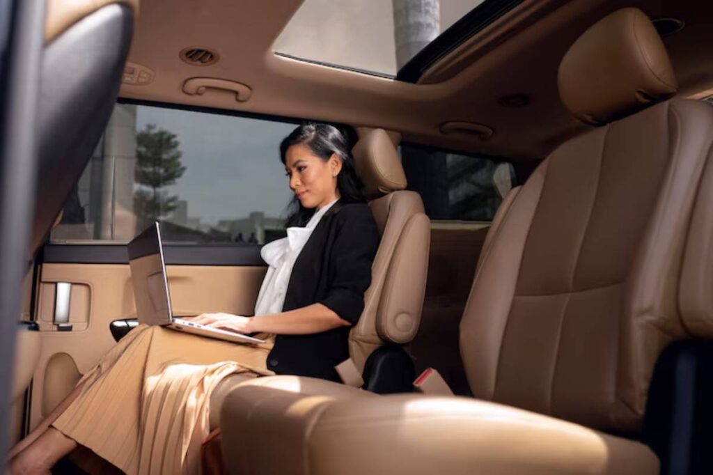 A professional woman in a car is working on a laptop, dressed in a black blazer and white blouse with a pleated skirt.