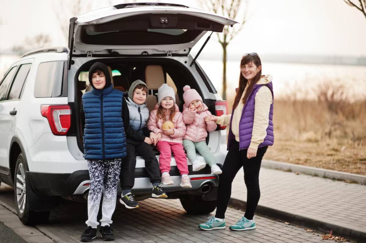 A group of children in colorful jackets sit on the back of an SUV in a scenic outdoor setting, enjoying snacks and each other's company.