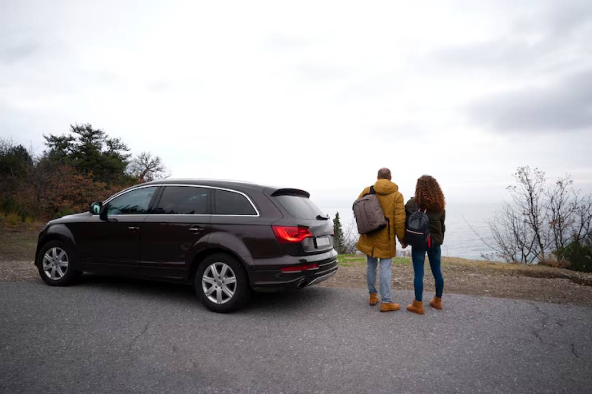 A couple stands hand in hand by a parked brown SUV, gazing at a scenic view of the ocean against a cloudy sky.