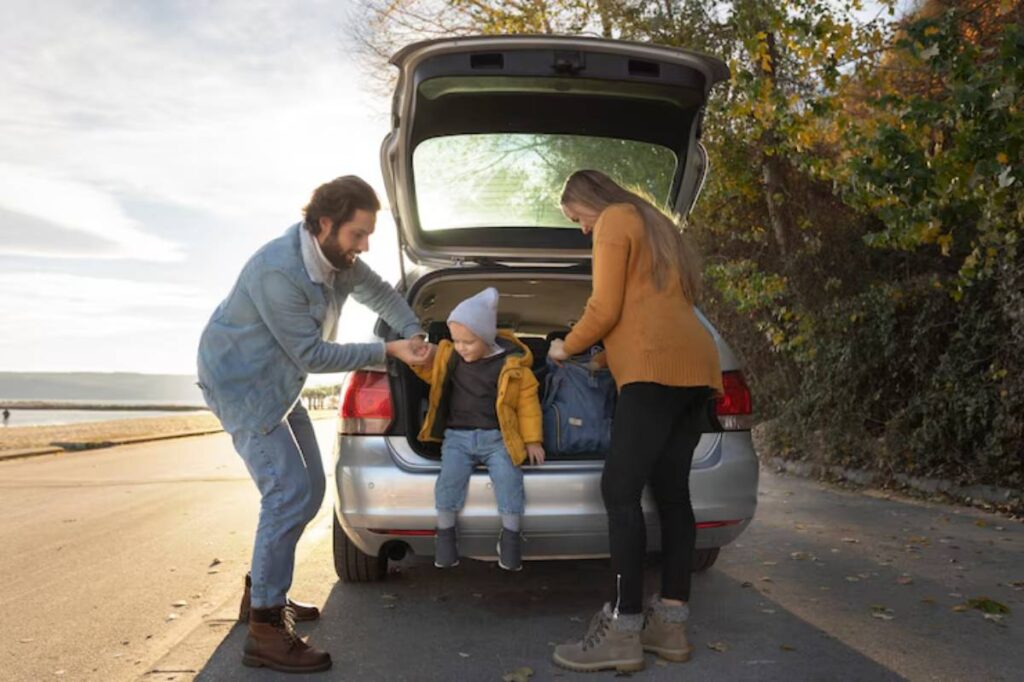 A family is unloading bags from the trunk of a car parked by a scenic waterfront during sunset, surrounded by autumn foliage.