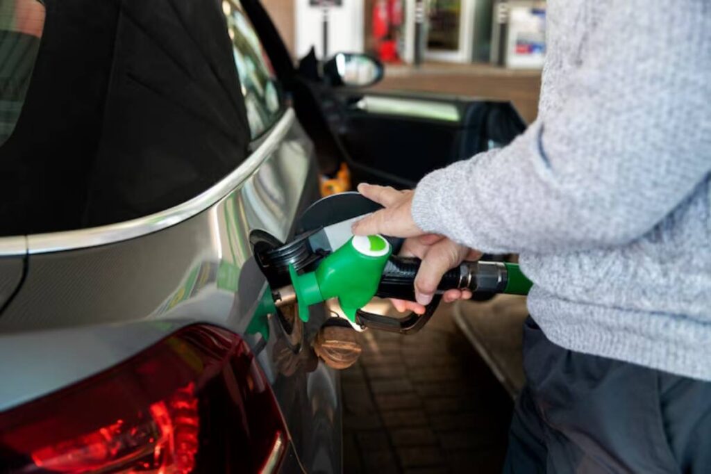 A person fills a silver car with fuel using a green gas pump nozzle at a gas station.