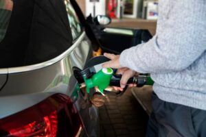 A person fills a silver car with fuel using a green gas pump nozzle at a gas station.