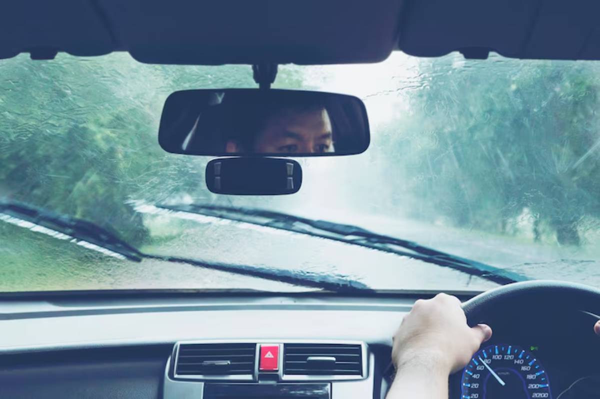 A driver's hands grip the steering wheel, while rain streaks down the windshield, obscuring the view of a winding road ahead.