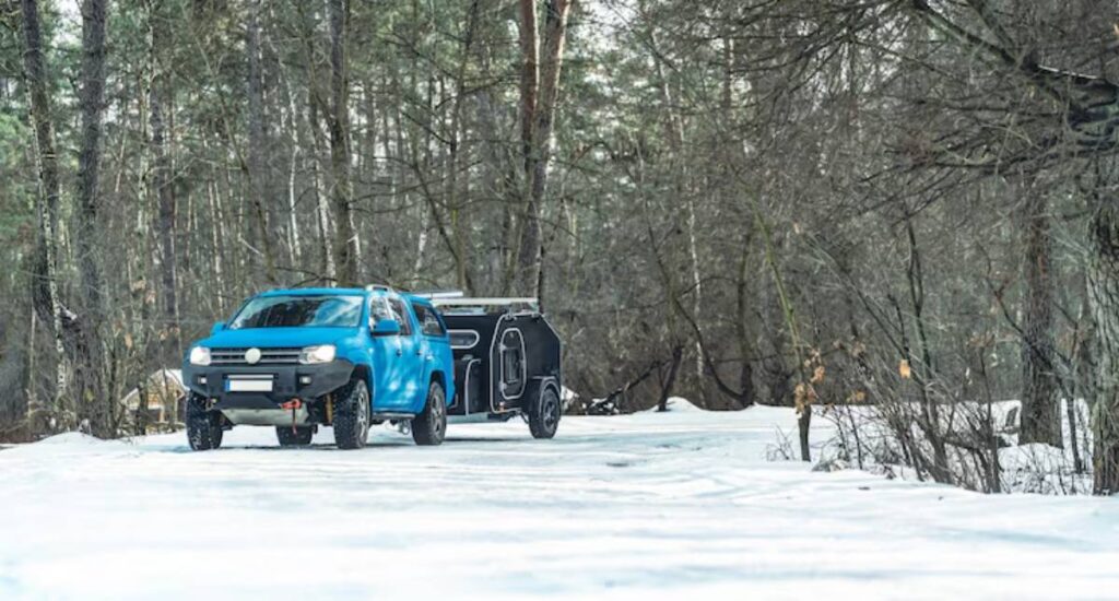 A blue off-road vehicle towing a compact black trailer drives on a snowy forest path surrounded by tall trees.
