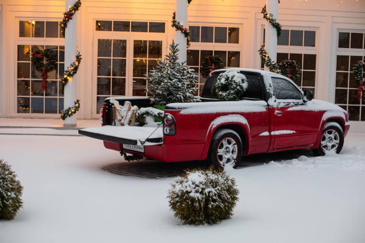 A red pickup truck covered in snow, parked in front of a festive house adorned with holiday decorations and greenery.