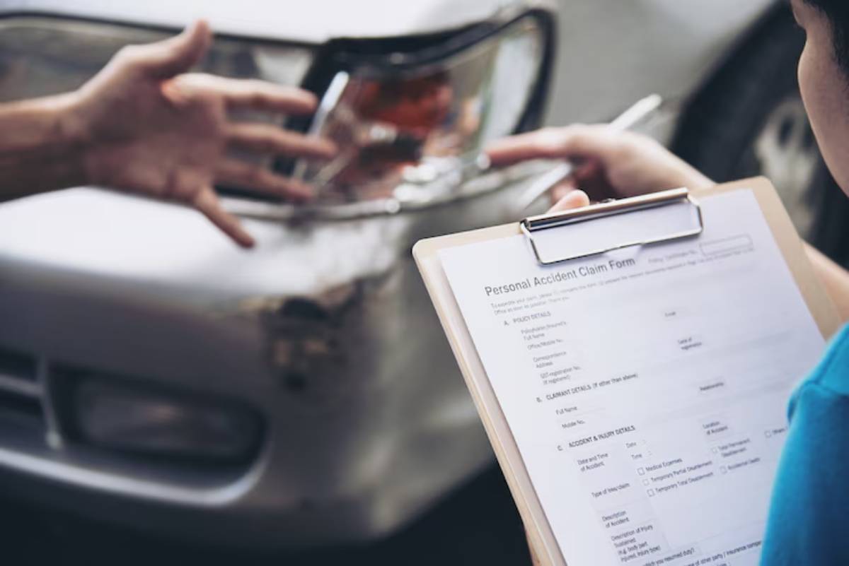 A person gestures towards a damaged vehicle while another holds a clipboard with a personal accident claim form.