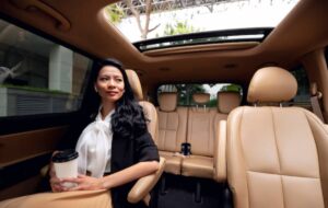 A woman in a black outfit and white blouse sits in a luxury car, holding a coffee cup, with spacious tan leather seats around her.