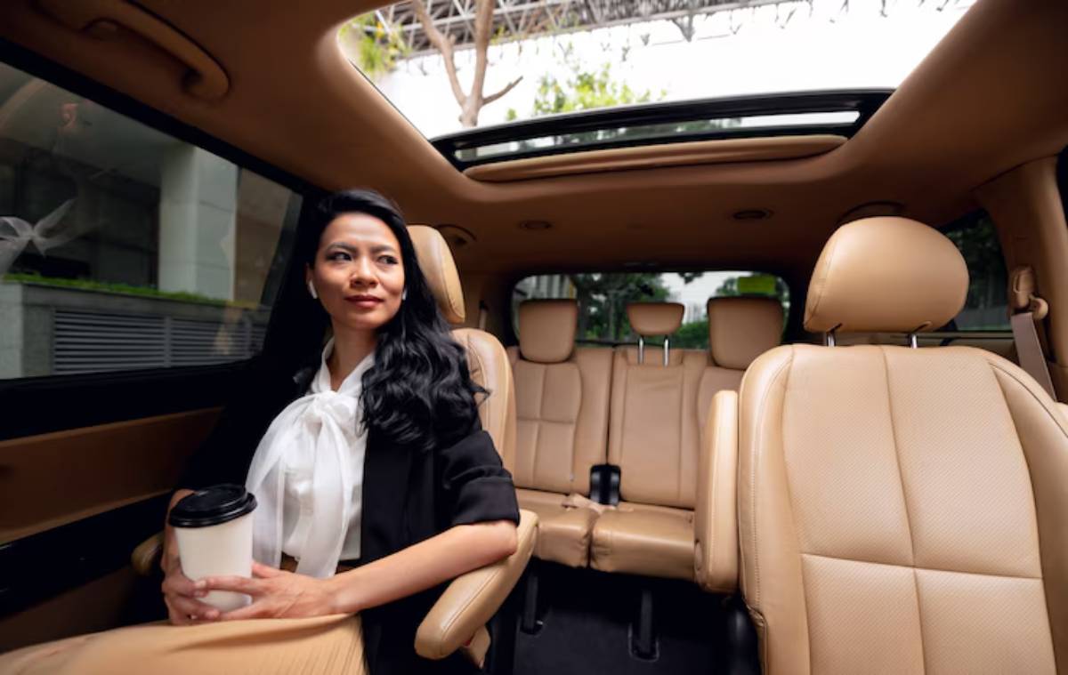 A woman in a black outfit and white blouse sits in a luxury car, holding a coffee cup, with spacious tan leather seats around her.