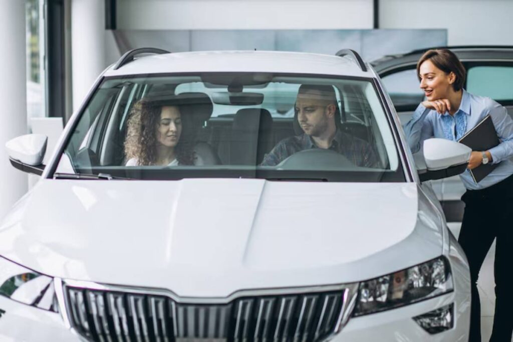 A sales representative discusses vehicle features with a couple seated inside a white car at a dealership.