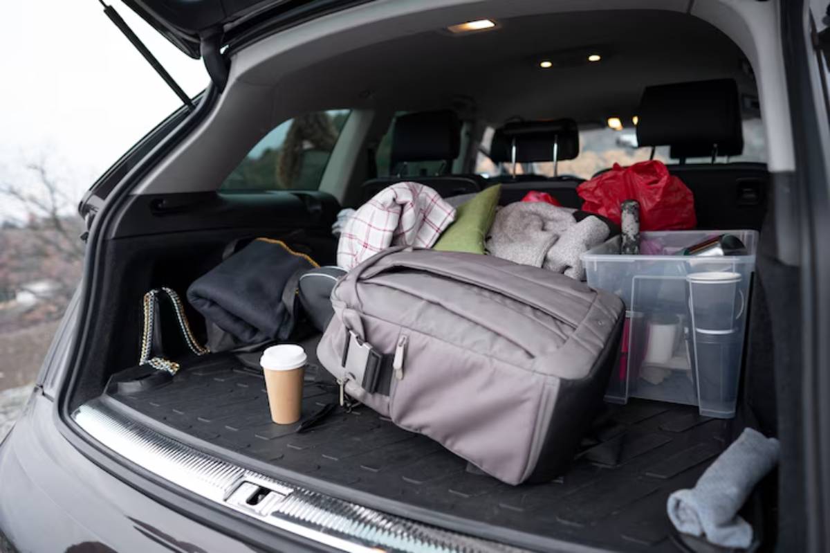 The back of a car is filled with clothing, bags, and storage containers, alongside a coffee cup on the floor.