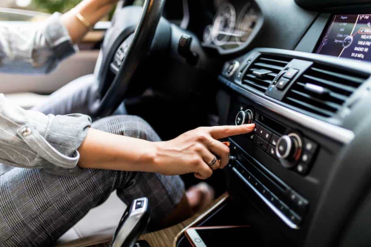 A person adjusts the car radio controls while driving, with a smartphone resting in the console.
