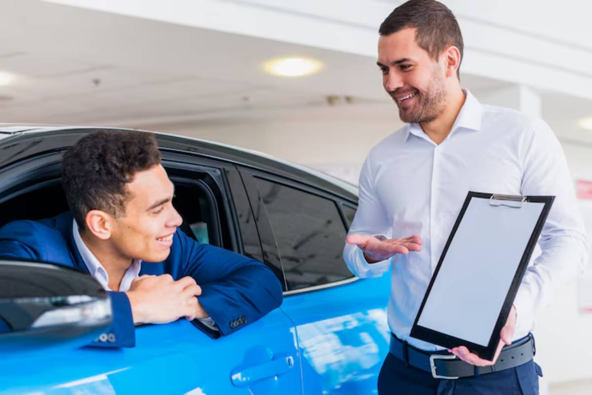 A car sales representative explains details to a prospective buyer leaning out of a blue car in a dealership setting.