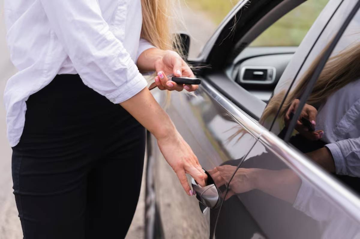 A woman in a white shirt uses a key fob to unlock a car door, standing beside a sleek gray vehicle on a rural road.