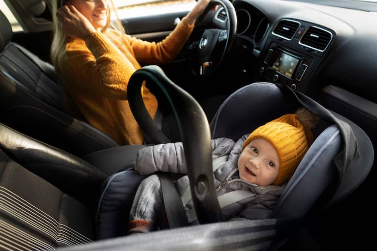 A woman wearing an orange sweater drives a car, while a child in a cozy, safety seat wears a yellow hat in the passenger area.