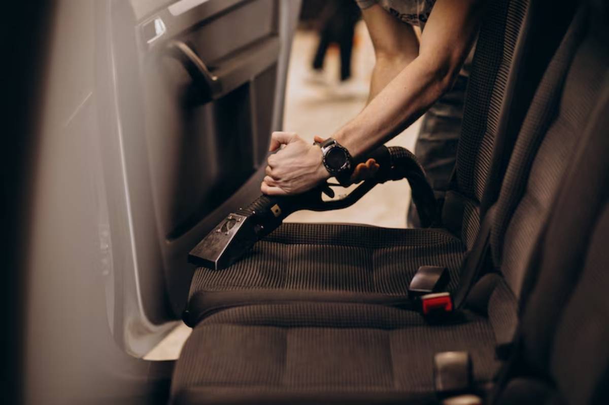 A person vacuums car seats with a handheld vacuum cleaner, focusing on the fabric upholstery.