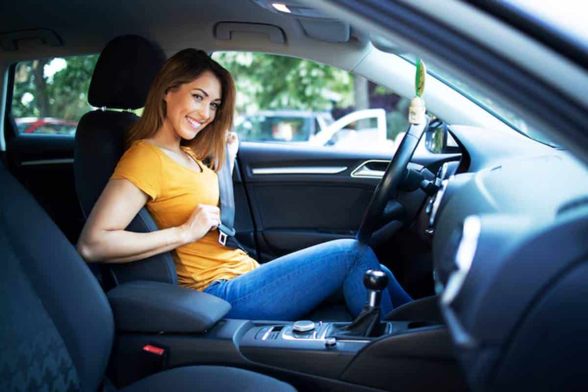 A woman in a yellow shirt is fastening her seatbelt while sitting in the driver's seat of a car, ready to drive.