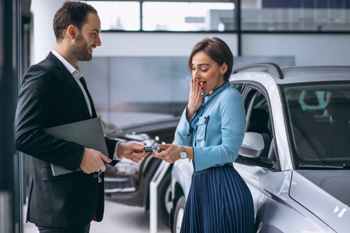 A suited man hands over keys to a woman in a blue blouse and skirt inside a modern car dealership, showcasing a vehicle in the background.