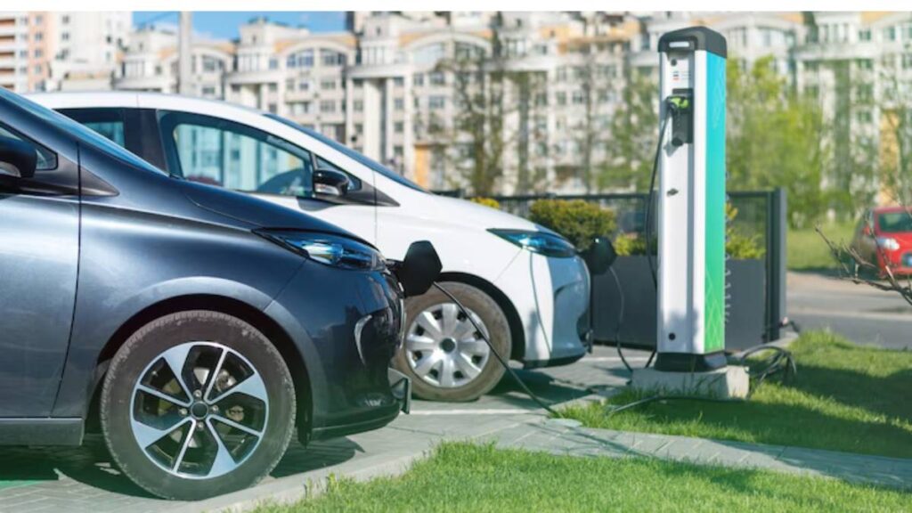 Two electric cars, a black one and a white one, are parked next to an electric vehicle charging station in a sunny urban setting.