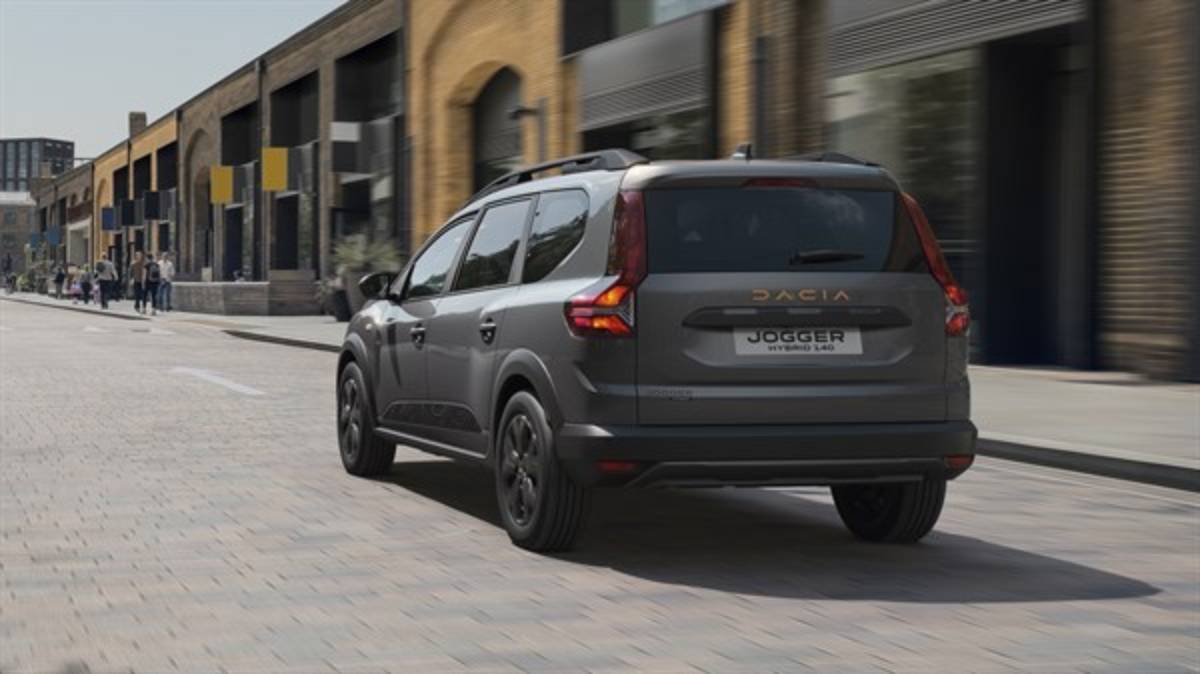 A sleek gray Dacia Jogger hybrid drives down a city street lined with modern buildings and pedestrians.
