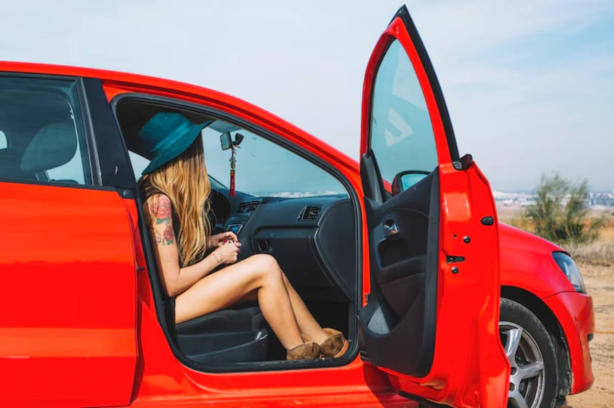 A woman with long hair and tattoos relaxes in a red car, wearing a blue hat, with the door open and a scenic landscape in the background.