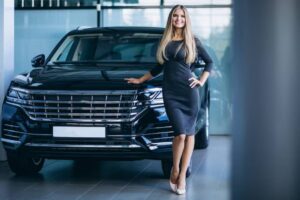 A stylish woman in a fitted black dress poses confidently next to a luxurious black SUV in a modern showroom.