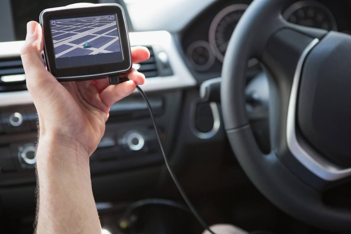 A hand holding a GPS device displaying a city map, with a car steering wheel visible in the background.