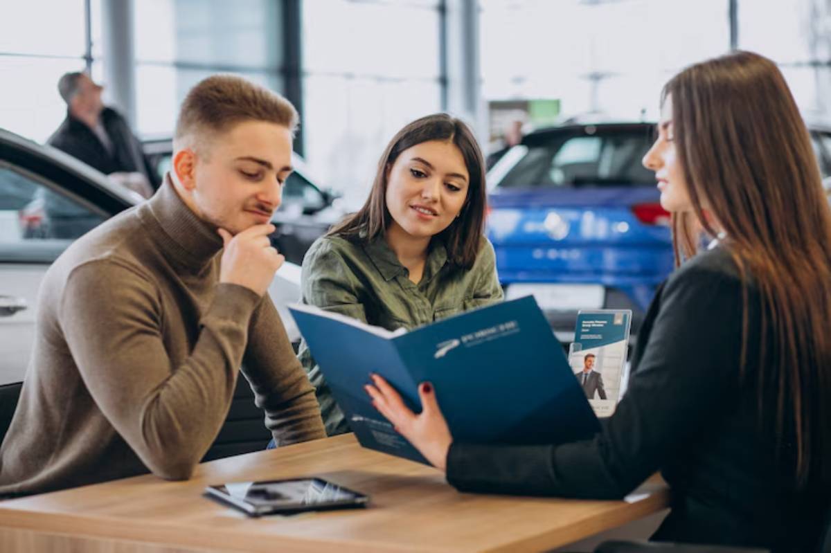 A customer listens attentively to a sales representative while reviewing a car brochure at a dealership. Cars are visible in the background.