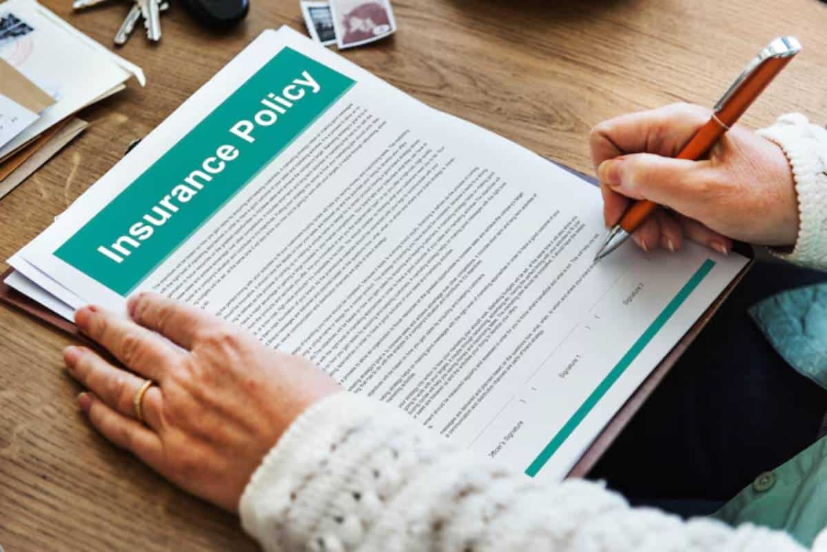 A person signing an insurance policy document on a wooden table, surrounded by a pen, keys, and some photos.