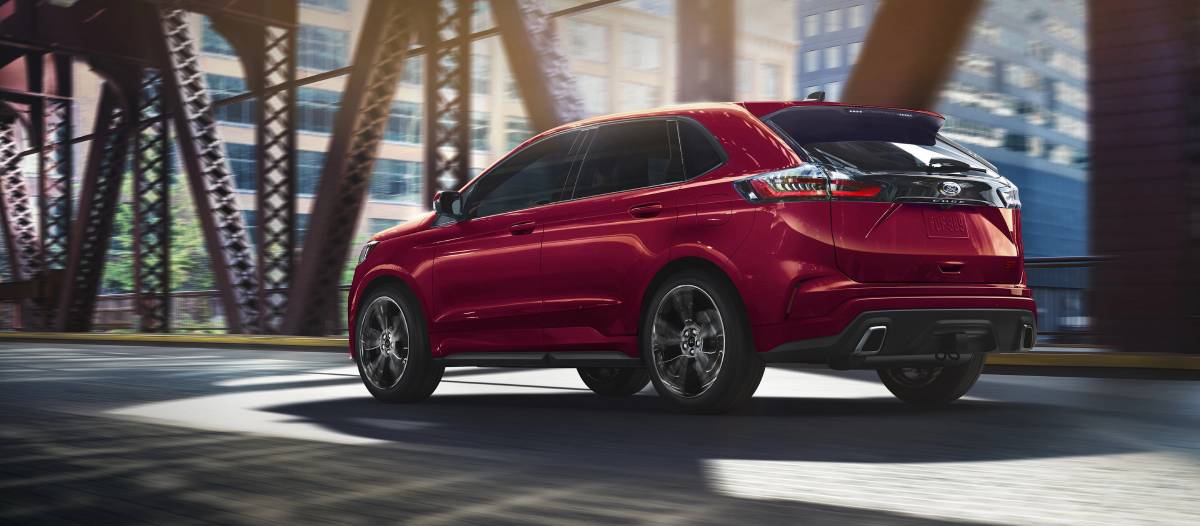 A sleek red SUV drives under a bridge in an urban setting, showcasing modern design and dynamic motion against a city backdrop.