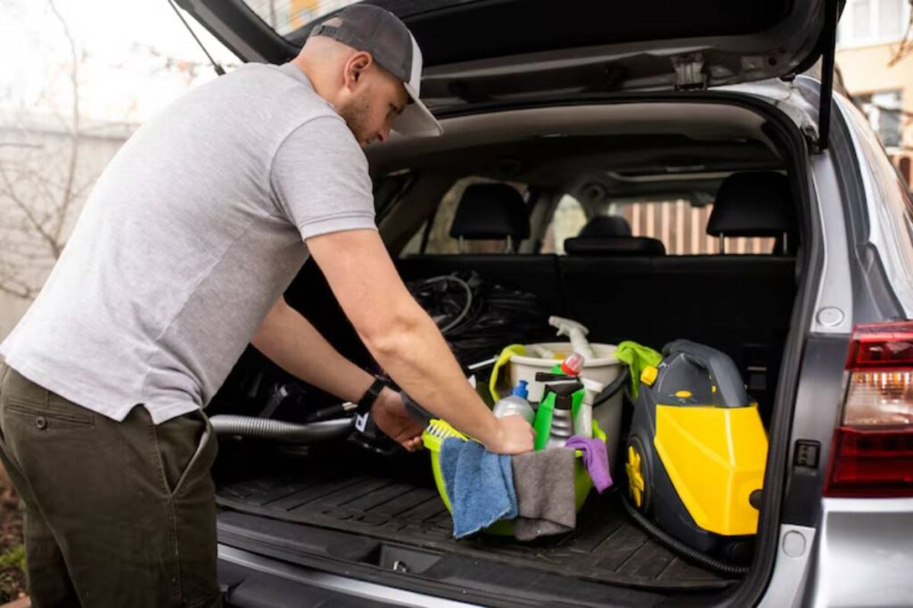 A person organizes cleaning supplies, including bottles and a vacuum, in the trunk of a silver SUV, surrounded by a natural setting.