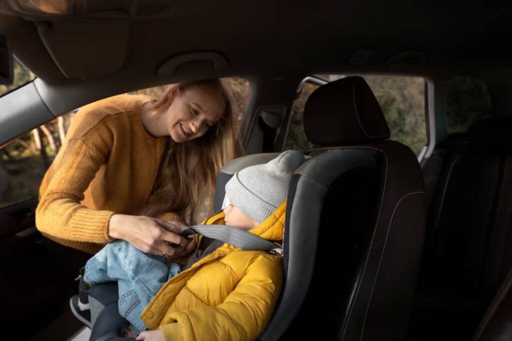 A child in a cozy yellow jacket and grey beanie is being buckled into a car seat by an adult, inside a vehicle.