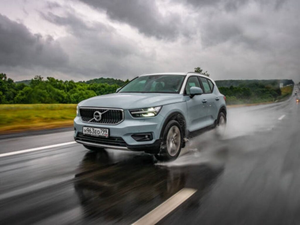 A blue Volvo SUV drives through rain-slicked roads, creating splashes, with dark clouds and greenery in the background.