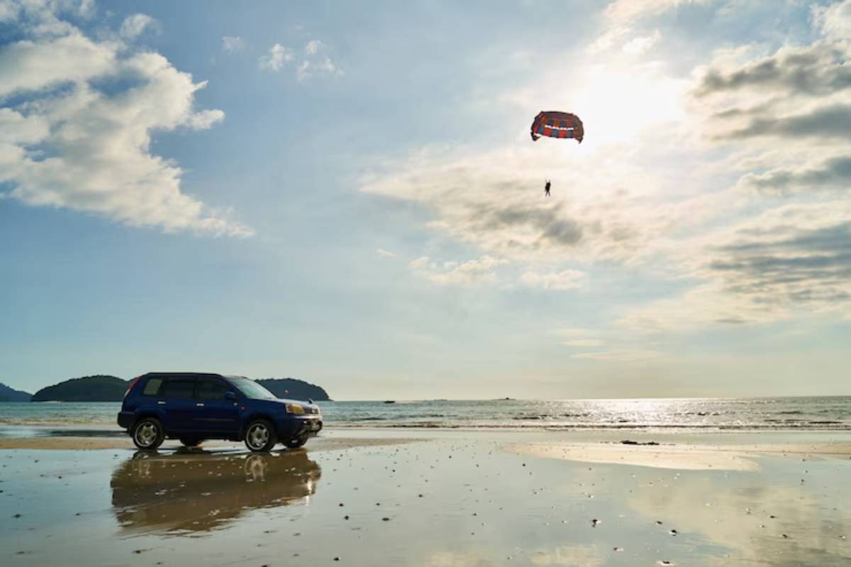 A blue SUV parked on a beach with a reflective wet surface, while a person paraglides above under a colorful parachute.