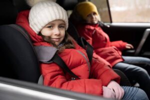 Two children in bright red jackets sit in a car, secured with seat belts, looking out the window on a chilly day.