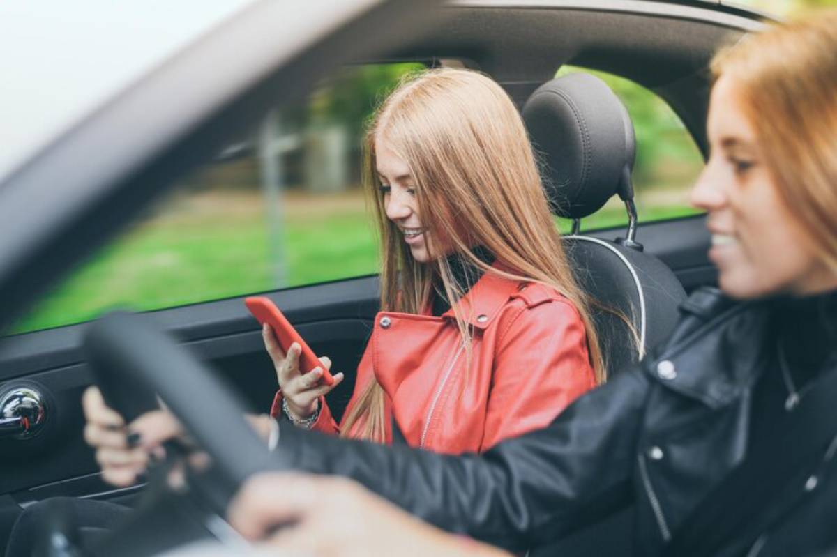 A young woman in a red leather jacket is looking at her phone while seated in a car, with a driver visible beside her.