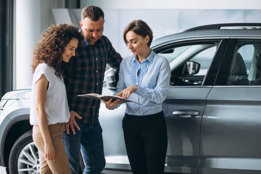 A woman in a light blue blouse shows a car brochure to a couple in a vehicle showroom, with a sleek gray car visible nearby.
