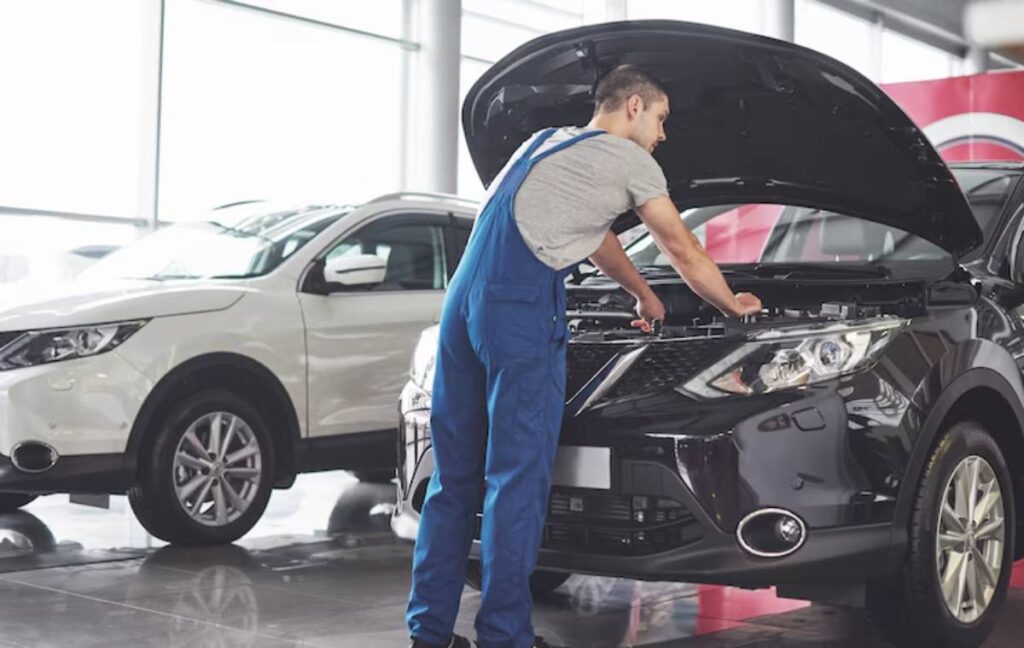 A mechanic in blue overalls inspects the engine of a black car in a bright showroom, with a white SUV visible in the background.