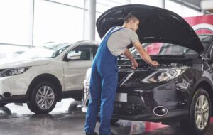 A mechanic in blue overalls inspects the engine of a black car in a bright showroom, with a white SUV visible in the background.