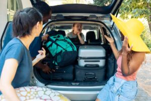 A group of friends packs a car with various luggage, including two suitcases and a green backpack, preparing for a trip.