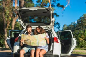 A couple sits in a car's trunk, studying a map against a backdrop of lush trees and a clear blue sky.