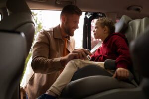 A man secures a child in a car seat, focused and attentive, with sunlight streaming through the open door.