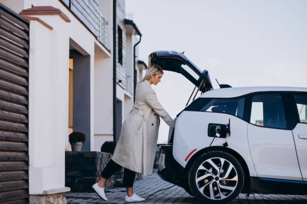 A woman in a long coat is loading items into the back of a white electric car parked in front of a modern house.