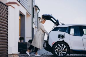 A woman in a long coat is loading items into the back of a white electric car parked in front of a modern house.