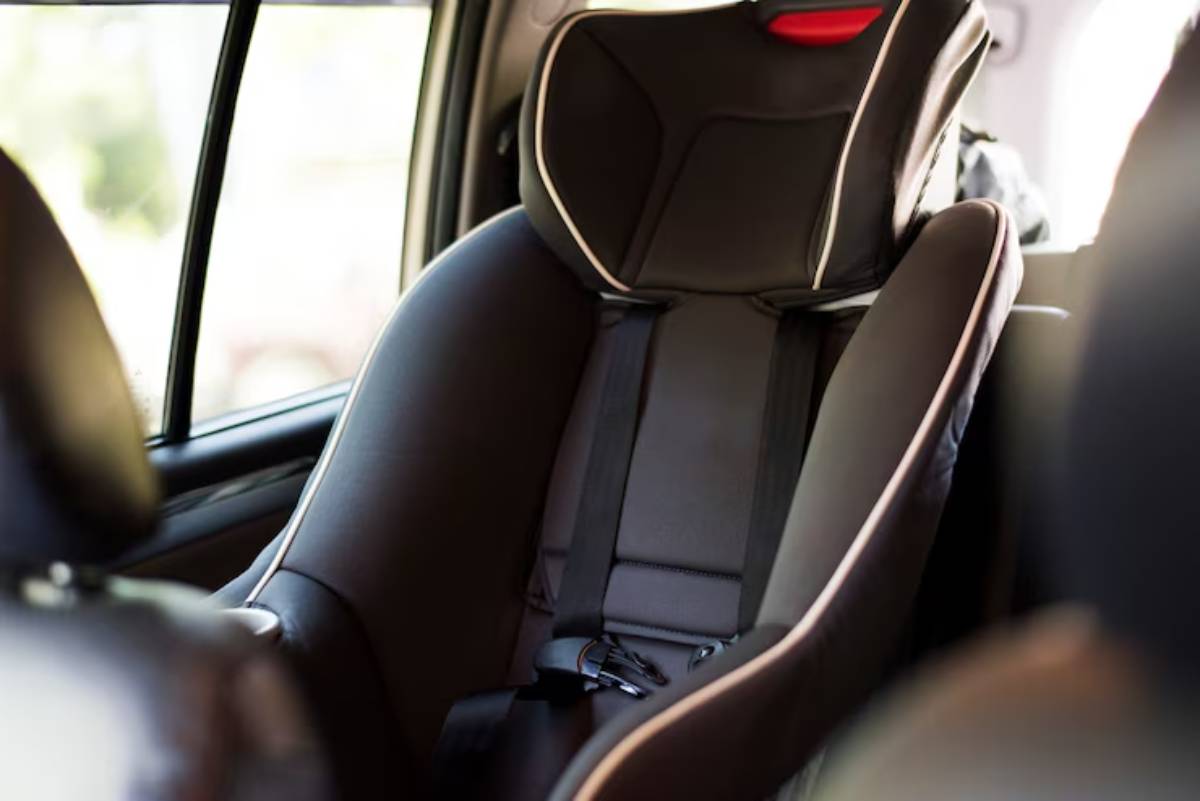 A black car seat with a high backrest and safety straps, positioned inside a vehicle, featuring natural light through the windows.