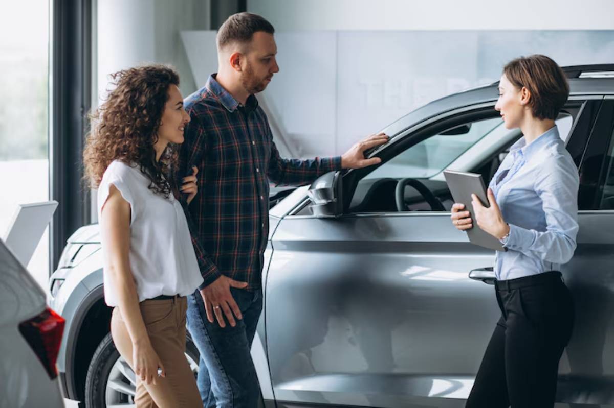 A couple discusses options with a salesperson next to a sleek gray car in a modern dealership, illuminated by natural light.