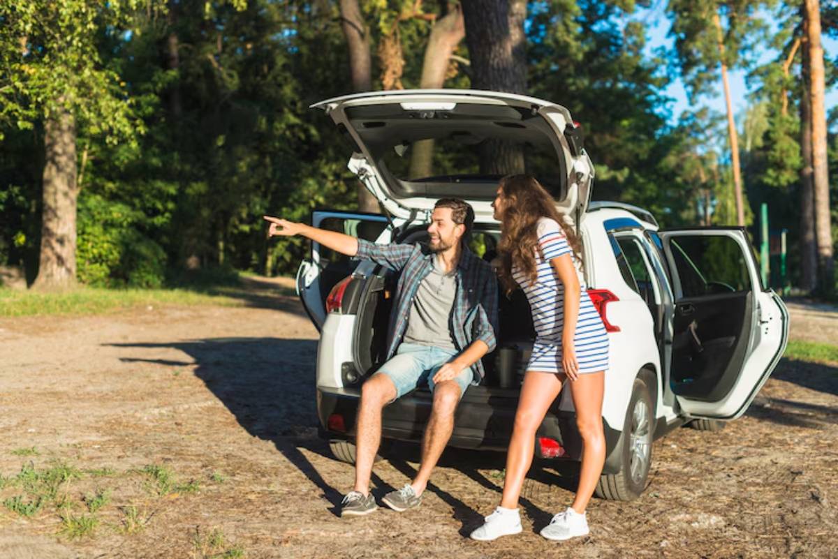A couple enjoys a sunny day, sitting by an open car trunk in a forested area, gesturing and chatting happily.