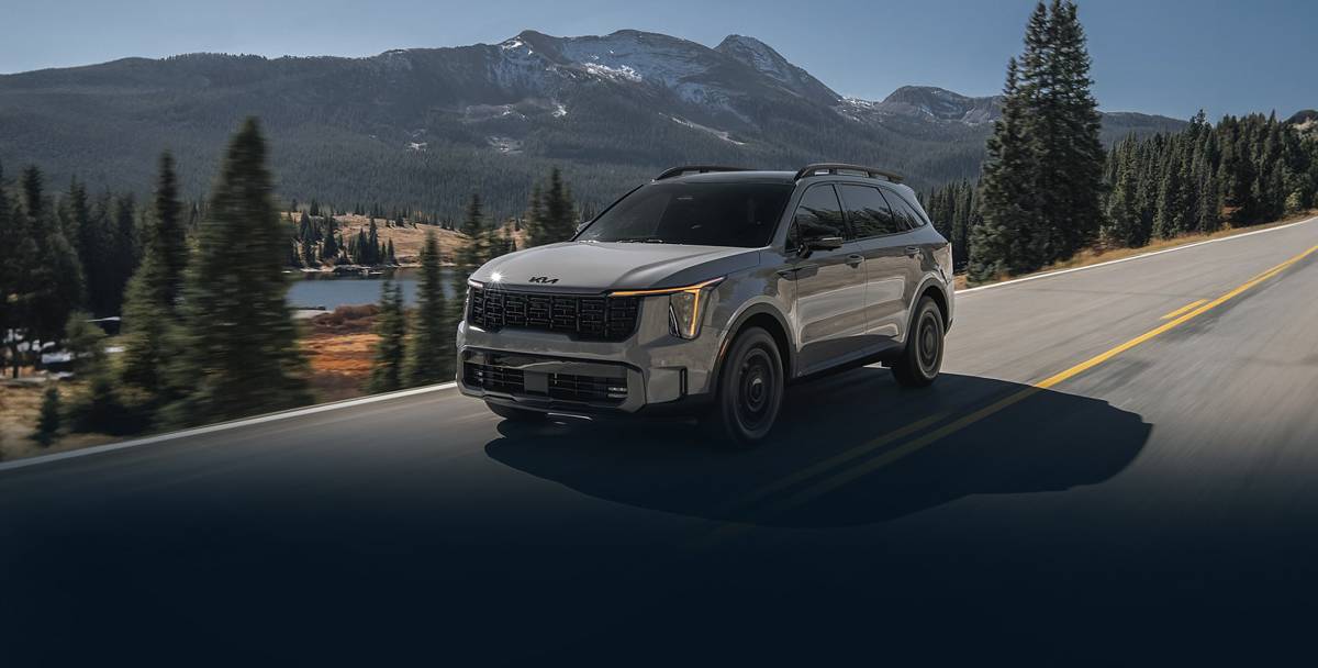 A modern SUV drives along a scenic mountain road, surrounded by lush trees and a reflective lake under a clear blue sky.